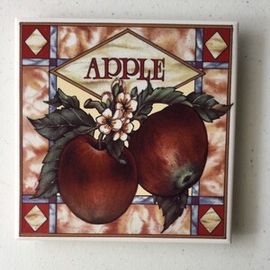 May include: A square ceramic tile with a stained glass design featuring two red apples with white blossoms and green leaves. The tile has the word "APPLE" in a yellow diamond shape at the top.