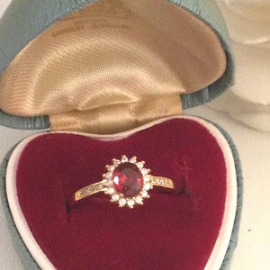 Può includere: Un anello d'oro con una grande gemma rossa ovale circondata da pietre bianche più piccole. L'anello è esposto in una scatola a forma di cuore, foderata in velluto rosso e con un esterno azzurro.