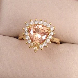 May include: A gold ring with a peach-coloured, triangular gemstone encircled by a halo of small, clear stones. The ring's band is plain, polished gold. The setting and halo create a sunburst effect around the central stone.