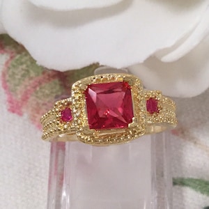 May include: A gold ring featuring a large square red gemstone, surrounded by a decorative gold border. Two smaller red gemstones are set on either side of the main stone.