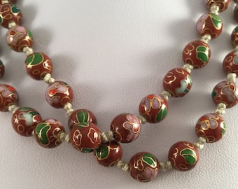 Puede incluir: Un collar de múltiples hilos hecho de cuentas redondas de color rojo con patrones florales en esmalte verde, rosa y blanco. Las cuentas están enhebradas en un cordón blanco.