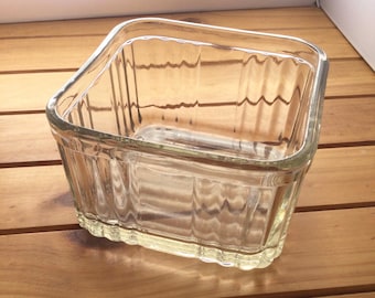 May include: A clear, rectangular glass baking dish with a ribbed design on the sides. The dish has a slightly rounded rim and sits on a wooden surface. The glass is transparent, allowing a view of the interior.