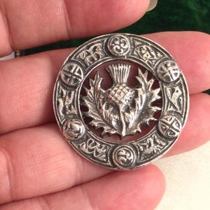 Puede incluir: Broche de plata con un diseño detallado de un cardo rodeado de nudos celtas. La broche es circular y tiene un acabado plateado.