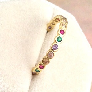 Puede incluir: Un anillo dorado con una serie de piedras preciosas de colores. Las piedras son redondas y están muy juntas, creando una banda continua de color. Las piedras incluyen tonos de rosa, morado, verde y transparente. El anillo es un accesorio de moda.