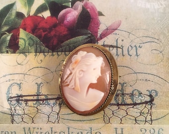 Peut inclure: Une broche camée représentant un profil de femme en camée blanc, sertie dans un cadre ovale ton or.