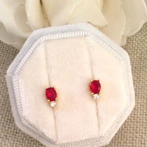 Può includere: Un paio di orecchini d'oro con pietre preziose ovali rosse e un singolo gioiello rotondo trasparente su ogni orecchino. Gli orecchini sono esposti in una scatola di velluto bianco.