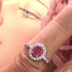 May include: A silver ring featuring an oval ruby gemstone, encircled by smaller, clear stones. The ring is displayed on a finger, with a soft-focus background.