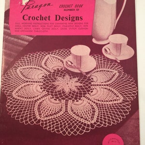 May include: A vintage crochet book cover featuring a close-up of a white crocheted doily with a delicate floral design. The book is titled "Paragon Crochet Book" and "Crochet Designs". The recommended price is 80 cents.