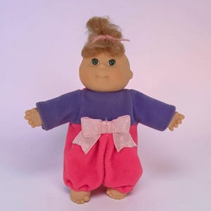 May include: A vintage doll with a soft, vinyl face and light brown hair styled in a bun. The doll wears a purple long-sleeved top and pink pants with a large pink bow. The doll's arms are outstretched.
