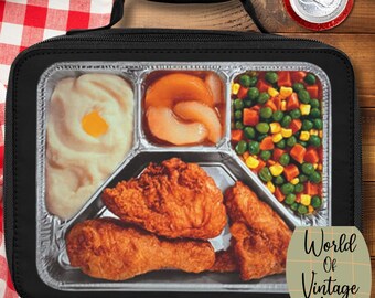 May include: Black lunchbox with a retro TV dinner design. The meal includes fried chicken, mashed potatoes, peaches, and mixed vegetables. A can of soda is visible in the upper right corner. The lunchbox is on a wooden surface with a red and white checkered cloth.