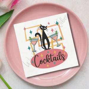 May include: A square white cocktail napkin with a black cat silhouette, two martini glasses, and the word "Cocktails" in a pink oval. The napkin is on a pink plate.