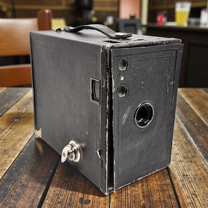 May include: A vintage black box camera with a leather handle and a silver-colored key. The camera has a rectangular shape with a lens and several circular openings on the front. It sits on a wooden surface.