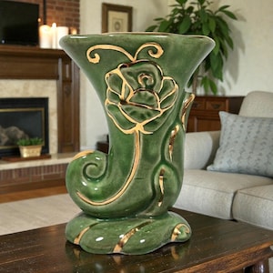 May include: Green ceramic vase with a gold rose design. The vase has a flared top and a curved, horn-like shape. Gold accents highlight the rose and the vase's edges. The vase sits on a round base and is a decorative home decor item.