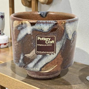 May include: A handcrafted stoneware Pottery Craft bowl with a brown, blue, and white marbled design. The bowl has a wide opening and a slightly flared base. The Pottery Craft label is visible on the front.