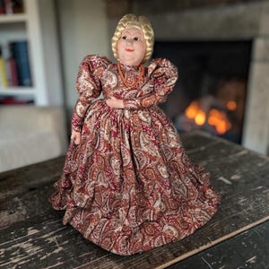 May include: A porcelain doll with blonde hair styled in braids, wearing a long-sleeved dress featuring a brown, red, and white paisley pattern. The doll has a painted face with red lips and wears a simple orange beaded necklace around its neck.