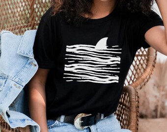 May include: A black t-shirt with a white graphic of a shark fin in the water. The graphic is made up of white stripes on a black background.