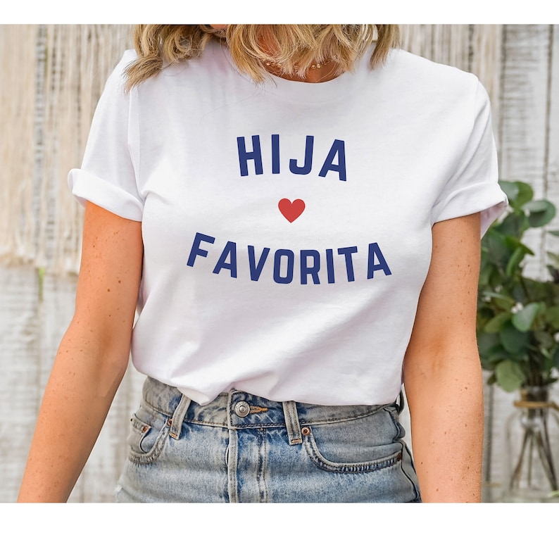 May include: White t-shirt with blue text that reads "HIJA" with a red heart and "FAVORITA" underneath.