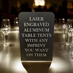 May include: A black sign with white text that reads "Laser Engraved Aluminum Table Tents With Any Imprint You Want On Them". The sign is sitting on a table with several clear glass wine glasses.