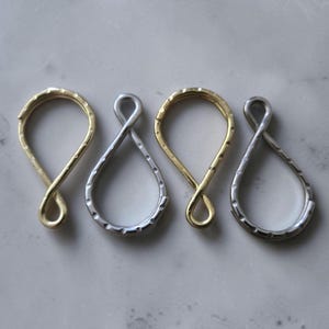 May include: Four teardrop-shaped metal clasps arranged in a row, alternating between gold-tone and silver-tone finishes. Each clasp features a twisted loop design at the bottom and a textured, hammered surface along the outer edge of the frame.