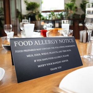 May include: A black sign with white text on a wooden table in a restaurant setting. The sign reads "Food Allergy Notice" and lists common allergens: milk, eggs, wheat, peanuts, and tree nuts. It also instructs guests with food allergies to notify their banquet server.