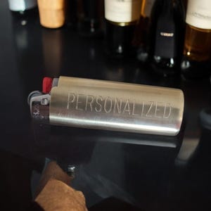 May include: A silver lighter with the word "PERSONALIZED" engraved on the side. The lighter is on a black surface.
