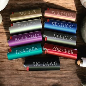 May include: Ten personalized lighters with various names and phrases engraved on them. The lighters are arranged in two rows of five, with the names facing the viewer. The lighters are all different colors, including gold, silver, blue, red, purple, green, and black.