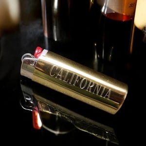 May include: A gold lighter with the word "CALIFORNIA" engraved on the side. The lighter is on a black surface.