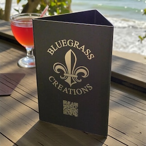 May include: A black triangular table tent with the words "Bluegrass Creations" and a fleur-de-lis logo in silver. A QR code is below the text. A red cocktail with a straw sits in a glass to the left. The background shows the ocean.