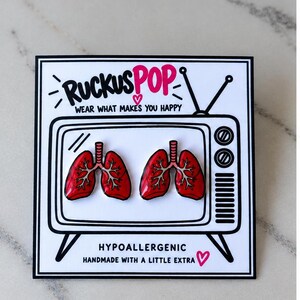 May include: A pair of red lung-shaped enamel stud earrings displayed on a white card featuring a black line drawing of a vintage television set with the text RUCKUSPOP WEAR WHAT MAKES YOU HAPPY and HYPOALLERGENIC HANDMADE WITH A LITTLE EXTRA.