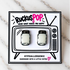 May include: A pair of stud earrings shaped like miniature mason jars with white centers, mounted on a white display card featuring a black and white illustration of a vintage television set with the text RUCKUSPOP WEAR WHAT MAKES YOU HAPPY.