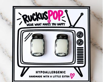 May include: A pair of stud earrings shaped like miniature mason jars with white centers, mounted on a white display card featuring a black and white illustration of a vintage television set with the text RUCKUSPOP WEAR WHAT MAKES YOU HAPPY.