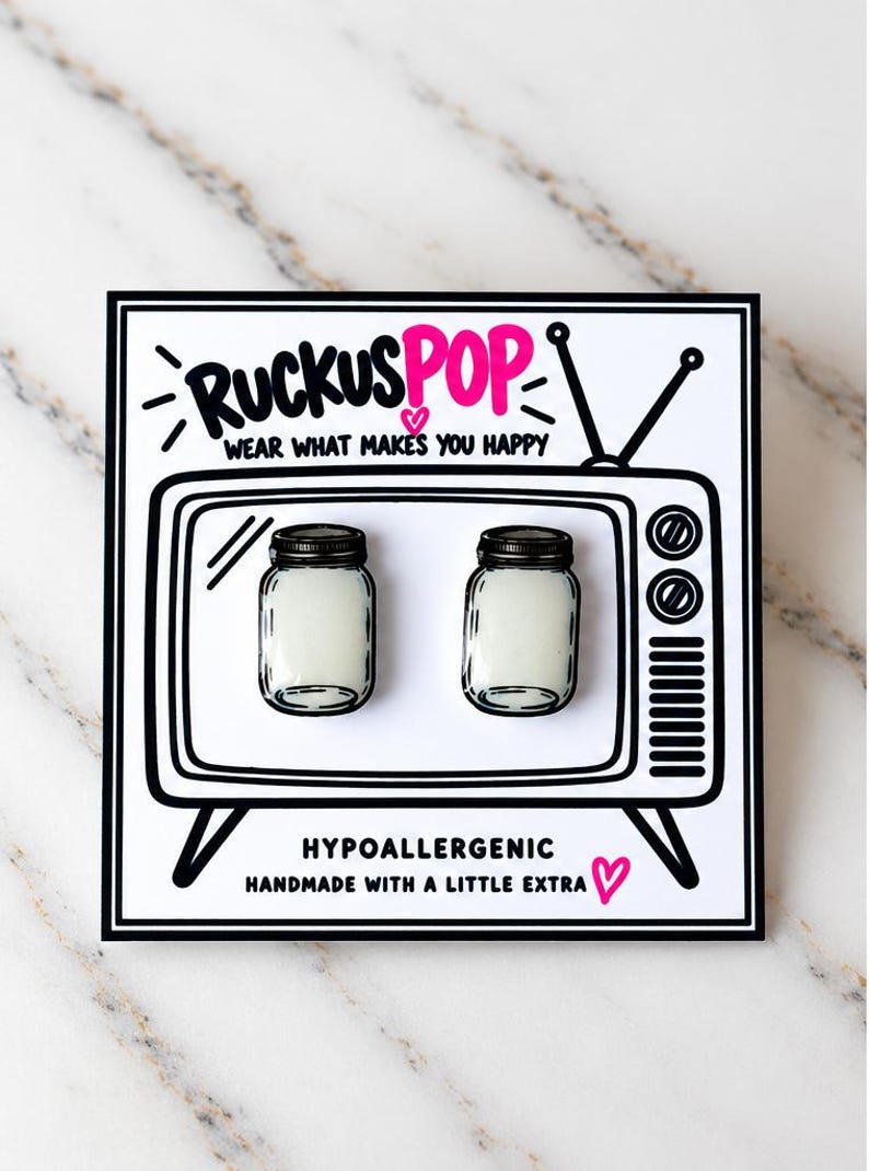 May include: A pair of stud earrings shaped like miniature mason jars with white centers, mounted on a white display card featuring a black and white illustration of a vintage television set with the text RUCKUSPOP WEAR WHAT MAKES YOU HAPPY.