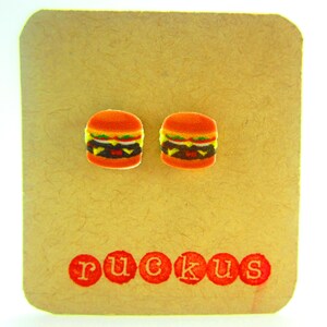 May include: A pair of small, colorful, hamburger-shaped earrings on a brown card with the word "ruckus" printed in red.