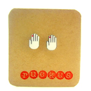 May include: A pair of white hand-shaped stud earrings with red nail polish on the index finger of each hand. The earrings are displayed on a brown card with the word "ruckus" printed in red.