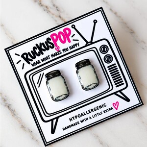 May include: A pair of stud earrings shaped like miniature mason jars with black lids and white contents, mounted on a white display card featuring a black line drawing of a vintage television and the text RUCKUSPOP WEAR WHAT MAKES YOU HAPPY.
