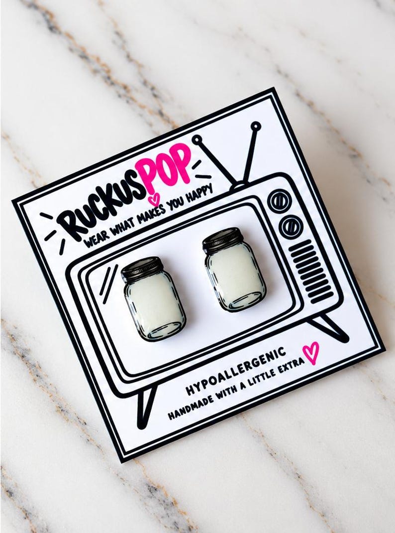 May include: A pair of stud earrings shaped like miniature mason jars with black lids and white contents, mounted on a white display card featuring a black line drawing of a vintage television and the text RUCKUSPOP WEAR WHAT MAKES YOU HAPPY.