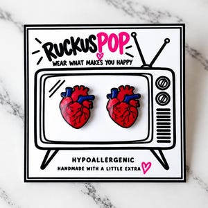 Pode incluir: Dois pins de esmalte em forma de cora&ccedil;&atilde;o anat&ocirc;mico com detalhes em vermelho e azul, apresentados num cart&atilde;o branco com o contorno de uma televis&atilde;o preta, o texto RUCKUSPOP, WEAR WHAT MAKES YOU HAPPY, HYPOALLERGENIC e HANDMADE WITH A LITTLE EXTRA.