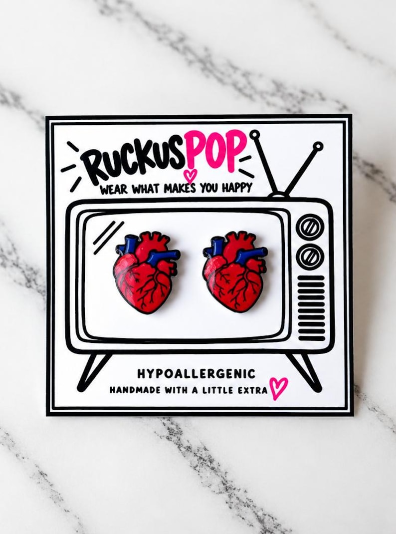 Pode incluir: Dois pins de esmalte em forma de cora&ccedil;&atilde;o anat&ocirc;mico com detalhes em vermelho e azul, apresentados num cart&atilde;o branco com o contorno de uma televis&atilde;o preta, o texto RUCKUSPOP, WEAR WHAT MAKES YOU HAPPY, HYPOALLERGENIC e HANDMADE WITH A LITTLE EXTRA.