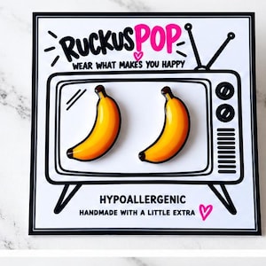 Pode incluir: Dois brincos amarelos em forma de banana exibidos num cartão branco com uma ilustração a preto e branco de uma televisão vintage com o texto RUCKUSPOP WEAR WHAT MAKES YOU HAPPY e HYPOALLERGENIC HANDMADE WITH A LITTLE EXTRA.