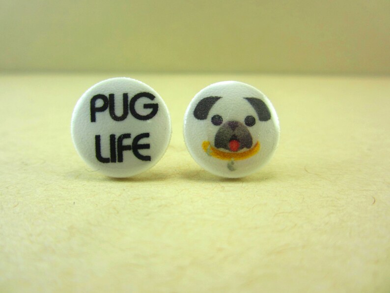 May include: A pair of white button earrings. One earring has a black and white pug face with a yellow banana. The other earring has the text "PUG LIFE" in black.