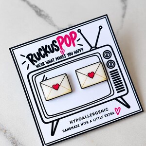 May include: Two envelope-shaped stud earrings with a cream background and a red heart in the center. They are attached to a white display card featuring a black line drawing of a vintage television and the text RUCKUSPOP WEAR WHAT MAKES YOU HAPPY.