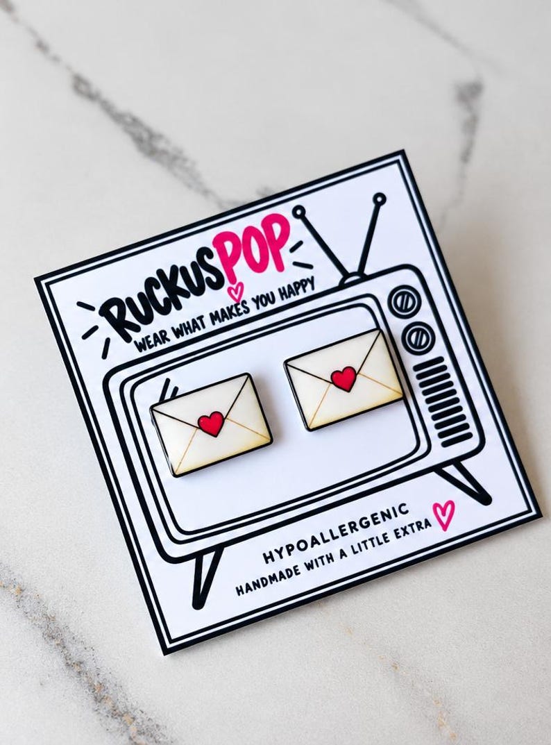 May include: Two envelope-shaped stud earrings with a cream background and a red heart in the center. They are attached to a white display card featuring a black line drawing of a vintage television and the text RUCKUSPOP WEAR WHAT MAKES YOU HAPPY.