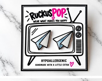 May include: Two white paper airplane enamel pins with blue accents displayed on a square backing card. The card features a black and white illustration of a vintage television set with the text RUCKUSPOP, WEAR WHAT MAKES YOU HAPPY, HYPOALLERGENIC, and HANDMADE WITH A LITTLE EXTRA.