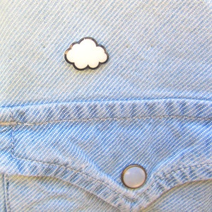 May include: A white cloud-shaped pin with a black outline, pinned to a blue denim shirt with a pocket and a button.