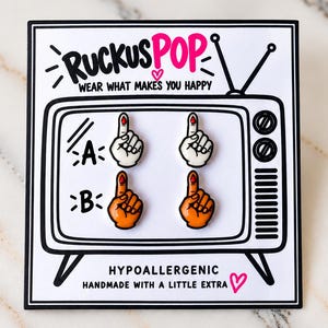 May include: Four enamel pins shaped like pointing fingers with red blood drops on the tips, arranged on a display card printed with a television graphic, the text RUCKUSPOP, WEAR WHAT MAKES YOU HAPPY, HYPOALLERGENIC, and HANDMADE WITH A LITTLE EXTRA.