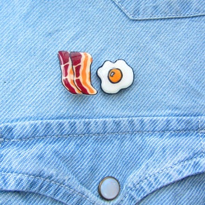 May include: Two ceramic pins on a blue denim background. One pin is shaped like a fried egg with a yellow yolk and white egg white. The other pin is shaped like two slices of bacon with red and orange stripes.