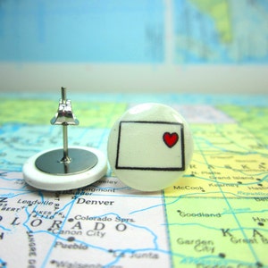 May include: A pair of white stud earrings with a black outline of the state of Colorado and a red heart in the center. The earrings are on a map of the United States.