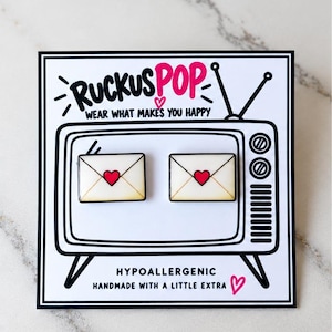 May include: Two rectangular enamel pins shaped like envelopes with a red heart in the center, displayed on a white card featuring a black and white illustration of a vintage television set with the text RUCKUS POP WEAR WHAT MAKES YOU HAPPY.