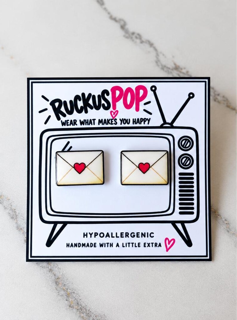 May include: Two rectangular enamel pins shaped like envelopes with a red heart in the center, displayed on a white card featuring a black and white illustration of a vintage television set with the text RUCKUS POP WEAR WHAT MAKES YOU HAPPY.