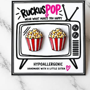 Pode incluir: Dois pins de esmalte em forma de baldes de pipoca com listras vermelhas e brancas em um cartão quadrado com uma ilustração em preto e branco de uma televisão vintage e o texto RUCKUSPOP WEAR WHAT MAKES YOU HAPPY HYPOALLERGENIC.